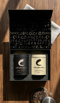 AI mock-up of gift box with 2 tea tins nestled in foam.  Box has Cs artwork printed on inside flap.  Tins are matte black and matte light yellow.