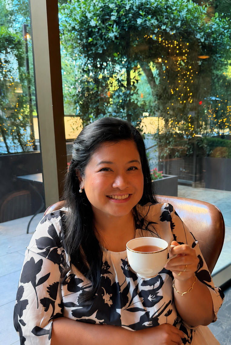 Photo of woman smiling and holding tea, view of outdoor patio in the window wall behind her.