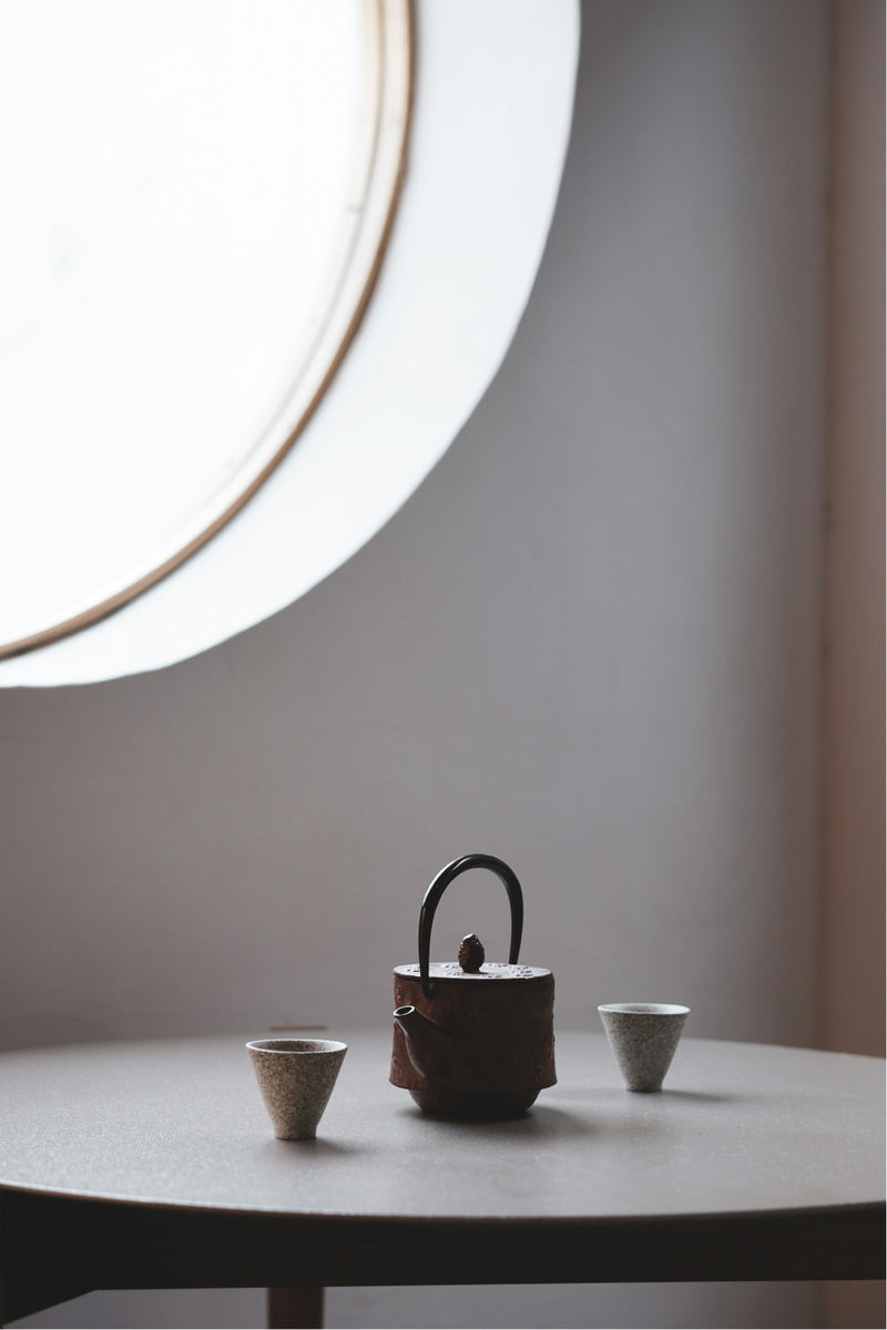 modern setting with large round window letting light in, simple table with small concrete looking teacups and a metal teapot
