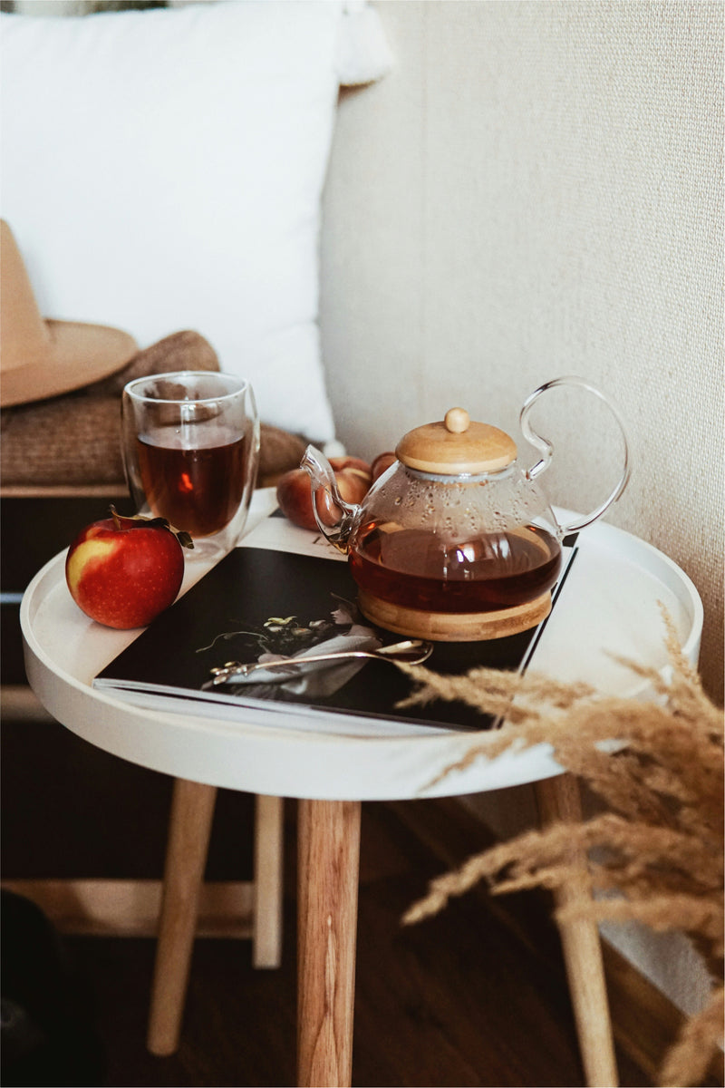 photo of very small table with a clear teapot, teacup, apple, and editorial magazine on top