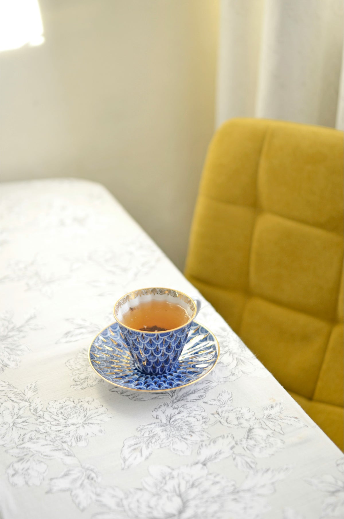 Photo of a teacup on a plate, both in a decorative blue, gold, and white pattern.  They are sitting on top of a light floral printed table cloth.